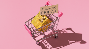 Metal shopping cart with black friday sign and golden gift box with hard shadow, on pink background.